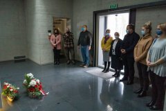 Uczestnicy wernisażu stoją przez kryptą w Pomniku Mauzoleum. Z lewej strony, na ziemi leżą kwiaty. W tle, za stojącymi wyjście na zewnątrz.