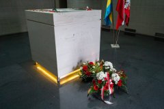 Krypta w Pomniku Mauzoleum. Na ziemi leżą kwiaty. W tle, w stojaku fragment flag: Polski, Mazowsza, Ostrołęki.