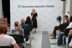 Kobieta przy mikrofonie przed widownią w tle napis Muzeum Kultury Kurpiowskiej 
