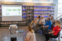 Uczestnicy edytonu zgromadzeni w bibliotece słuchają prelekcji. W tle ekran z wyświetloną prezentacją