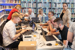 Uczestnicy edytonu przy wspólnym stole pracują przy komputerach. W tle regały biblioteczne