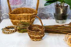 Na stole mały koszyk z wikliny, w tle duży koszyk z wikliny, obok ozdoby z wikliny w kształcie kółek oraz fragment pęku wikliny