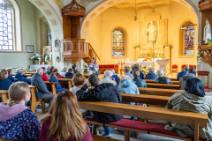Uczestnicy wewnątrz kościoła słuchają przemówienia prowadzącej, w tle ołtarz, ambona i fragment lewej nawy bocznej