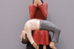 Rzeźba siedzącej, długowłosej postaci w czerwonym stroju, z dłońmi zakrywającymi twarz. Postać ma kolanach na osobę leżącą na plecach ze zwieszoną do tyłu głową, zwieszonymi dłońmi i nogami.