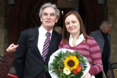 Starszy mężczyzna pozuje do zdjęcia z kobietą trzymającą bukiet kwiatów. W tle inne osoby.