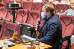 Mężczyzna w maseczce z mikrofonem w dłoni siedzi przy stoliku, widok z boku. Na stoliku otwarty laptop, mikrofon oraz okablowanie. Z lewej strony kadru stoi projektor multimedialny. W tle fragment widowni z publicznością.
