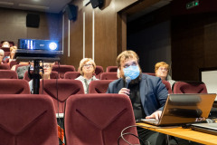 Mężczyzna w maseczce z mikrofonem w dłoni siedzi przy stoliku, widok od przodu. Na stoliku laptopy, mikrofon, okablowanie. Z lewej strony kadru stoi projektor multimedialny. W tle fragment widowni z publicznością.