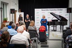 Fragment widowni, w tle trzy kobiety stoją  przy mikrofonach, fortepian i ścianka z logami