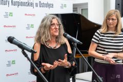 Dwie kobiety stoją, jedna z nich przemawia do mikrofonu. W tle ścianka z logami