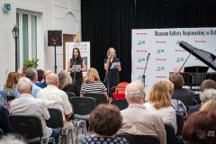 Fragment widowni, w tle dwie kobiety stoją  przy mikrofonach, fortepian i ścianka z logami