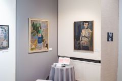 Fragment wystawy dzieł malarskich, stolik z materiałami promocyjnymi wystawy, na ścianach obrazy