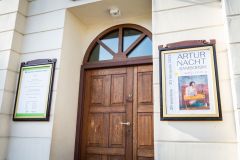 Wejście do muzeum, po bokach którego wiszą: po lewej tablica z informacją o godzinach otwarcia, po prawej plakat wystawy "Artur Nacht-Samborski. Wejrzenia"