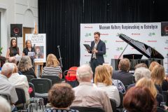 Fragment widowni, w tle dwie kobiety i mężczyzna stoją  przy mikrofonach, fortepian i ścianka z logami