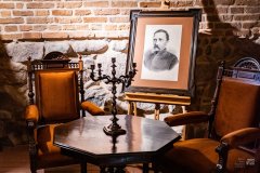 Zabytkowy stoliki krzesła w muzealnej piwnicy. Za stolikiem stoi sztaluga ze czarno-białym portretem mężczyzny
