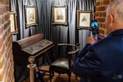 Na szarej zasłonie wiszą czarno-białe ilustracje w ramach przedstawiające portrety mężczyzn. Poniżej, na ziemi stoi zabytkowe krzesło i sekretarzyk. Z prawej strony fragment mężczyzny robiącego smartfonem zdjęcie ekspozycji.