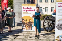 Na zewnątrz przy starej armacie stoi kobieta, przemawia do mikrofonu, obok fragment planszowej wystawy plenerowej, w tle budynek, z lewej strony kadru stoi pięć osób