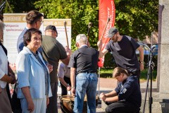 Na zewnątrz kilku mężczyzn uderza pałkami w instrument perkusyjny, jeden z mężczyzn kuca napinając membranę, obok stoi przyglądający się im mężczyzna oraz kobieta