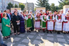 Kobiety i mężczyźni stoją w większości w strojach ludowych, w tle budynki