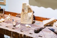 Fragment wystawy, warsztat rzeźbiarza na którym stoi klocek drewna z naszkicowaną postacią, na etapie tworzenia rzeźby