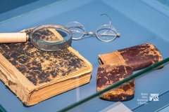W szklanej gablocie stara podniszczona książka, na niej leży lupa, obok książki okulary i etui