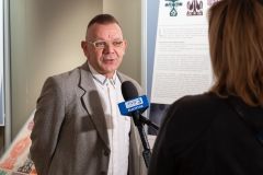 Mężczyzna udziela wywiadu telewizji. W tle fragment wystawy.