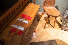 Drewniana, zdobiona ławka na której leżą niedokończona wycinanki. Na podłodze przy ławce leżą skrawki papieru powstałe podczas wycinania. Obok stoi drewniane zabytkowe zdobione krzesło.