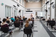 Kobieta stoi, przemawia do mikrofonu. Przed nią fragment widowni. W tle ścianka z logotypami oraz napisem Muzeum Kultury Kurpiowskiej w Ostrołęce.