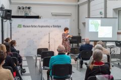 Kobieta prezentuje plansze na ekranie projektora. Przed nią fragment widowni. W tle ścianka z logotypami oraz napisem Muzeum Kultury Kurpiowskiej w Ostrołęce.