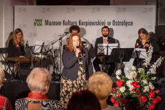 Kobieta z mikrofonem stoi przed sceną. W tle, na scenie siedzą muzycy. Przez sceną kilka osób z widowni.