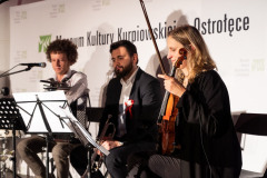Troje muzyków siedząc gra na scenie.