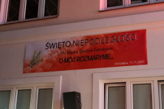 Na ścianie biało-czerwony banner z napisami: Święto Niepodległości  XIV wspólne śpiewanie patriotyczne O mój rozmarynie Ostrołęka 11.11.2021. Pod bannerem i nad nim okna.