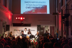 Chłopiec w kapeluszu typu cylinder, z mikrofonem w dłoni stoi na scenie. Za nim siedzą muzycy. Nad sceną wyświetlony biało-czerwony banner z napisem Święto Niepodległości XIV Wspólne Śpiewanie Patriotyczne O mój rozmarynie Ostrołęka 11.11.2021. Przez sceną kilka osób z widowni.