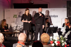 Mężczyzna i kobieta z mikrofonami stoją na scenie. Mężczyzna śpiewa. Z nimi siedzą muzycy. Z prawej strony sceny biało-czerwony bukiet kwiatów. Przed sceną kilka osób z widowni.