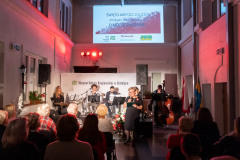 Kobieta z mikrofonem stoi obok sceny. Przed nią fragment widowni. Za nią, na scenie muzycy. Nad sceną, wyświetlony biało-czerwony banner z napisami Święto Niepodległości XIV wspólne śpiewanie patriotyczne O mój rozmarynie, na dole bannera logotypy: Muzeum Kultury Kurpiowskiej, marki Mazowsze oraz Powiatu Ostrołęckiego