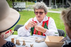 Twórczyni w stroju kurpiowskim siedząc przy stole wykonuje byśki. Po drugiej stronie stołu siedzą jeszcze dwie osoby.