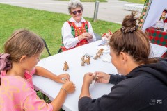 W plenerze przy stole siedzi twórczyni w stroju kurpiowskim i wykonuje byśki. Po drugiej stronie stołu siedzi kobieta oraz dziewczynka, uczące się wykonywania pieczywa obrzędowego.