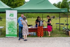 Trzy kobiety stoją przy stole pod namiotem typu pawilon. Przed namiotem, obok rollupu Muzeum Kultury Kurpiowskiej stoi kobieta i mężczyzna. W tle za namiotem zieleń.