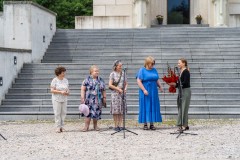 Pięć kobiet stoi przy schodach Pomnika Mauzoleum. Jedna z nich wręcza drugiej bukiet kwiatów.