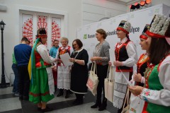 Na muzealnym dziedzińcu stoi 8 kobiet, w tym 5 w kurpiowskich strojach ludowych. W tle ścianka napisem Muzeum Kultury Kurpiowskiej w Ostrołęce i logami. Z prawej strony fragment choinki świątecznej.