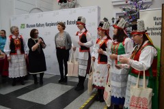 Na muzealnym dziedzińcu stoi 8 kobiet, w tym 5 w kurpiowskich strojach ludowych. W tle ścianka napisem Muzeum Kultury Kurpiowskiej w Ostrołęce i logami. Z prawej strony fragment choinki świątecznej.