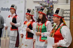 Na muzealnym dziedzińcu stoi 4 kobiety w kurpiowskich strojach ludowych. W tle ścianka napisem Muzeum Kultury Kurpiowskiej w Ostrołęce i logami. Z prawej strony fragment choinki świątecznej.