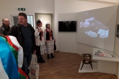 Dwóch mężczyzn oraz dwie kobiety stoją na wystawie przed ekranem, na którym wyświetlany jest monochromatyczny film. Kobiety są w stroju kurpiowskim.