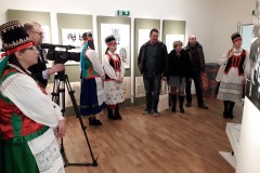 Cztery kobiety w stroju kurpiowskim oraz dwóch mężczyzn i kobieta w stroju codziennym stoją na wystawie wycinanki. Jeden z mężczyzna 