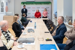 Kilka osób siedzących przy długim stole patrzy w stronę trzech osób stojących. W tle ścianka z logami marki Mazowsze i MKK