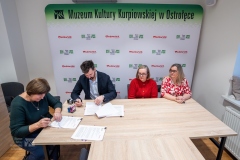 Cztery osoby siedzące przy jednym stole, mężczyzna z kobietą podpisują dokumenty. W tle ścianka z logami marki Mazowsze i MKK