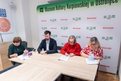 Cztery osoby siedzące przy jednym stole, trzy kobiety podpisują dokumenty. W tle ścianka z logami marki Mazowsze i MKK