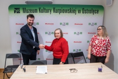 Kobieta z mężczyzną podający sobie rękę w zawarciu umowy, po prawej stoi druga kobieta spoglądająca w ich stronę. W tle ścianka z logami marki Mazowsze i MKK